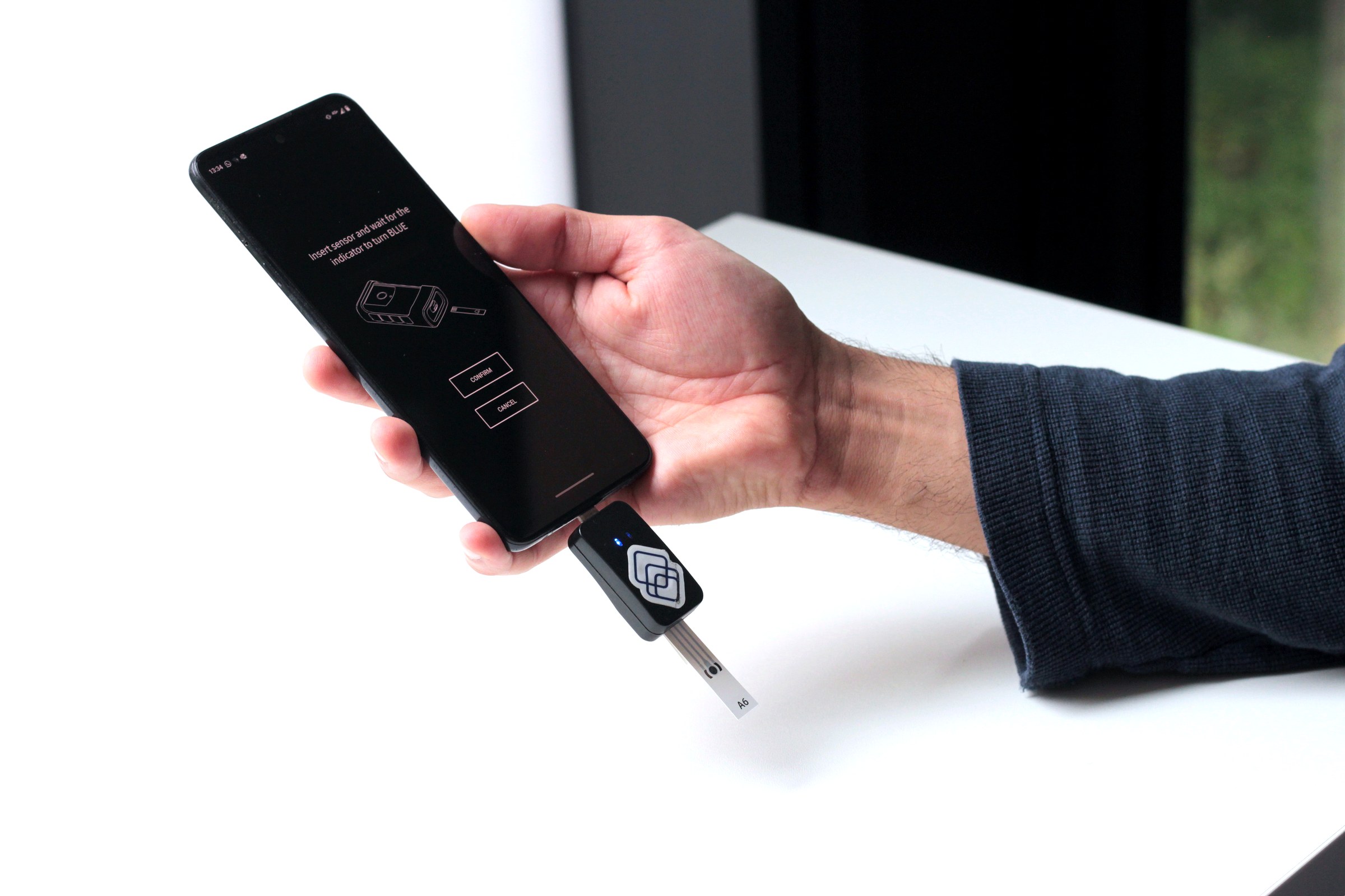 Hand holding the Segura reader connected to a smartphone showing the app